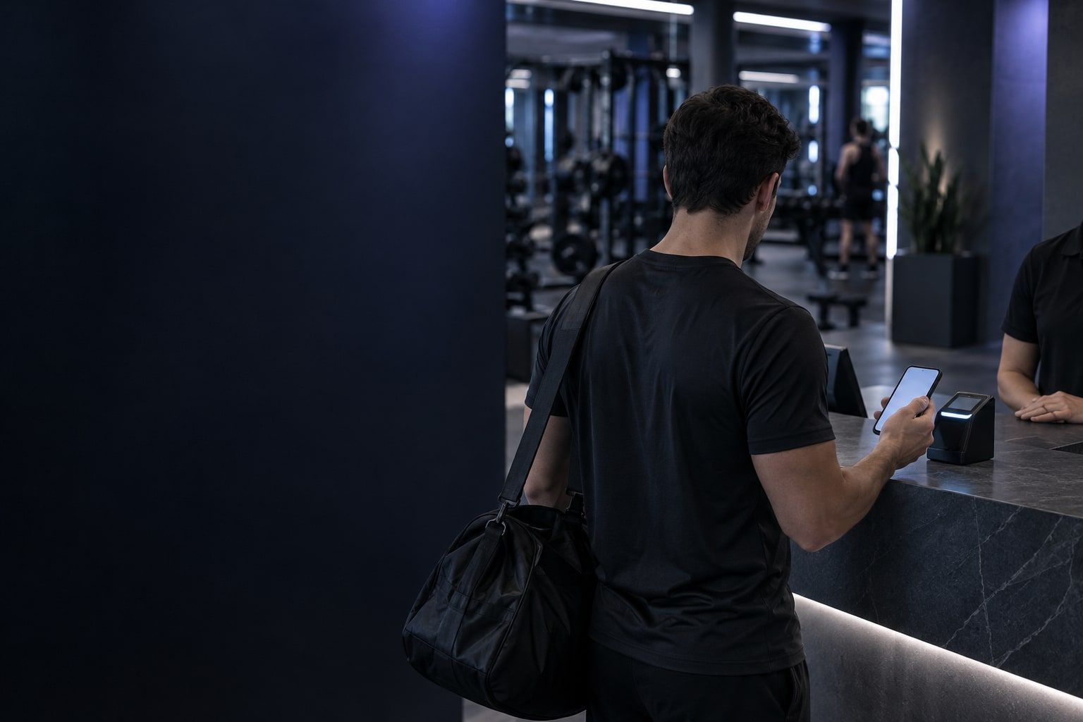 A man, seen from behind, holds his phone to the scanner at a dark stone front desk, with staff behind the counter.